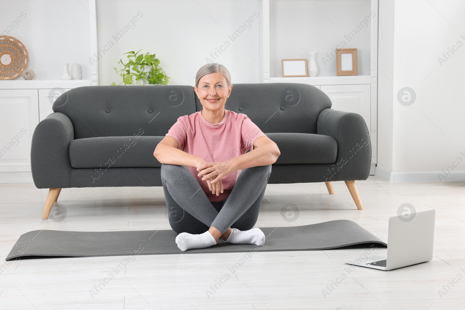 Photo of Senior woman in sportswear sitting on fitness mat near laptop at home