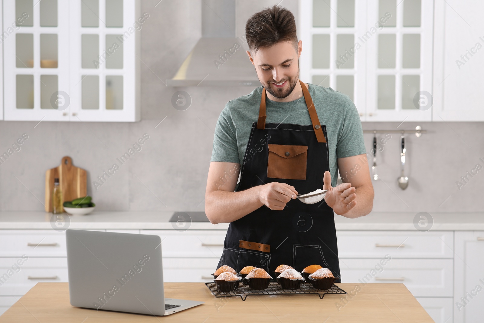 Photo of Man decorating muffins with powdered sugar while watching online cooking course via laptop in kitchen. Time for hobby