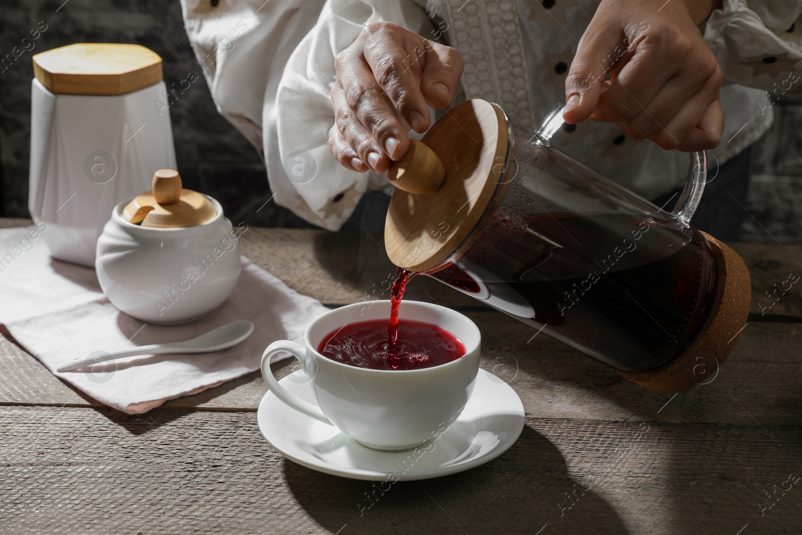 Photo of Woman pouring freshly brewed hibiscus tea from teapot into cup at wooden table, closeup