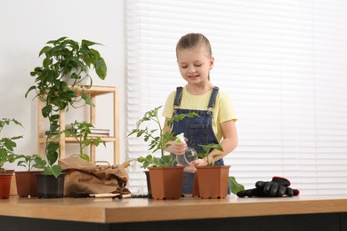 Photo of Cute little girl spraying seedling in pot at wooden table in room
