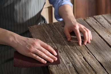 Photo of Man polishing wooden table with sandpaper indoors, closeup
