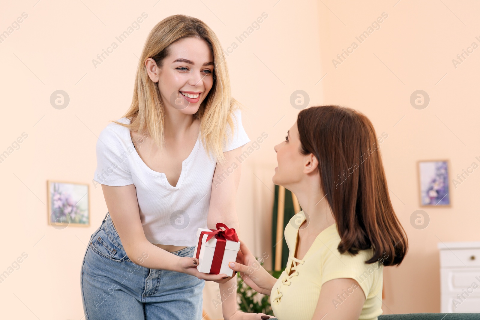 Photo of Smiling young woman presenting gift to her friend at home