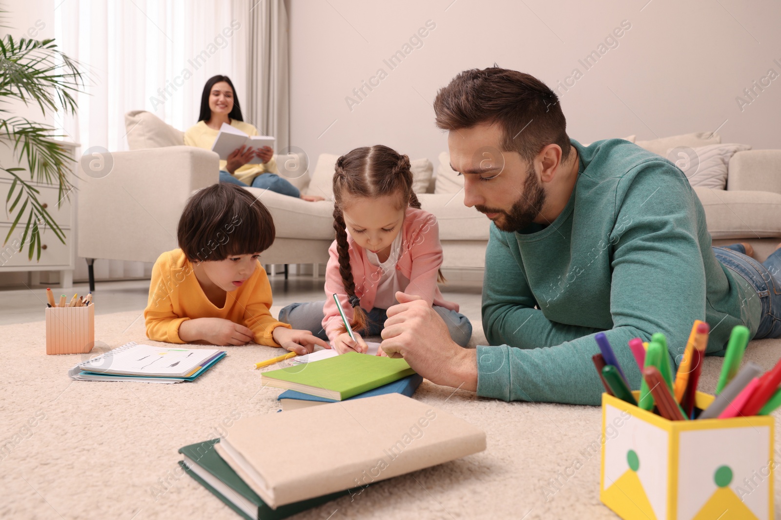 Photo of Father playing with his children while mother reading book on sofa in living room