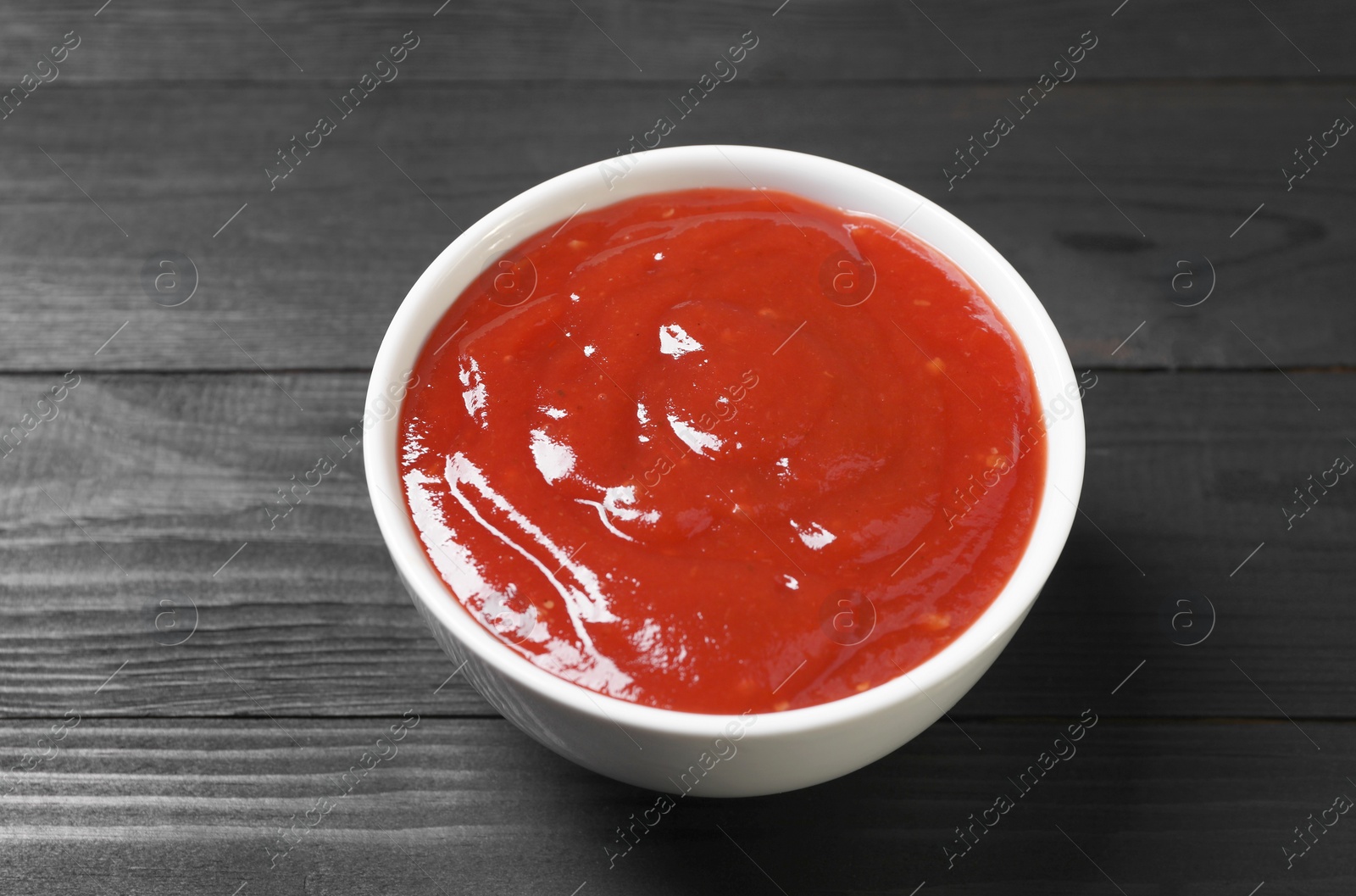 Photo of Delicious ketchup in bowl on black wooden table, closeup. Tomato sauce