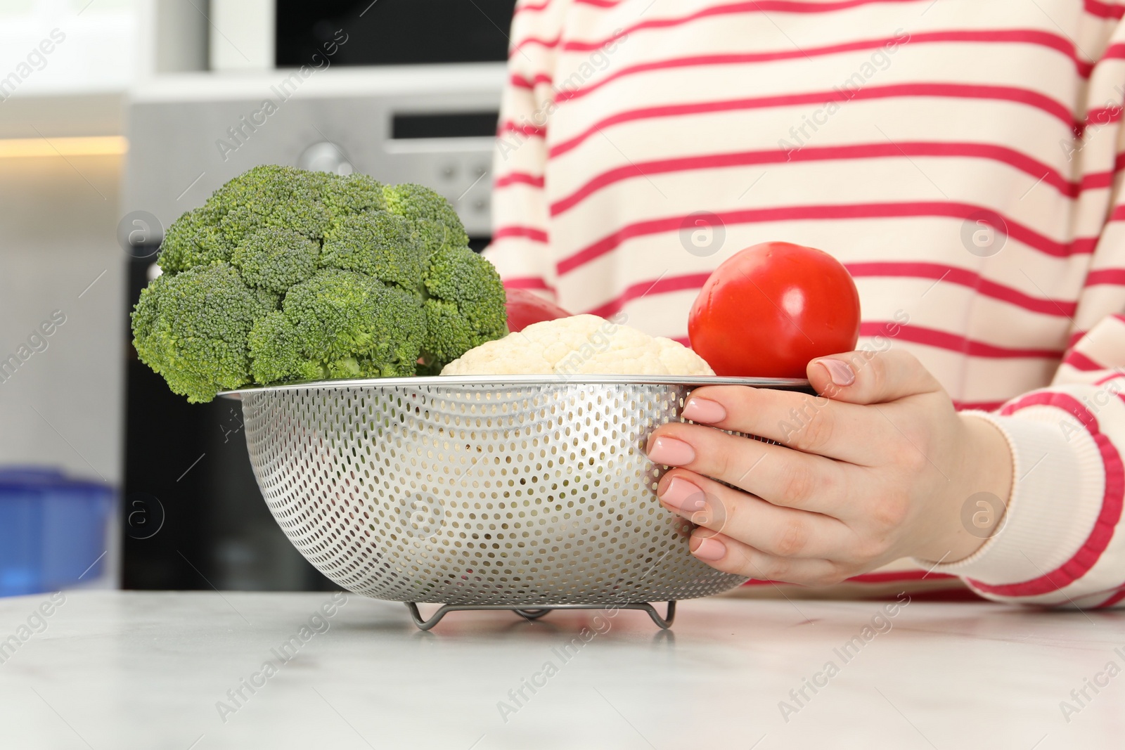 Photo of Woman holding colander with fresh vegetables at white marble table in kitchen, closeup