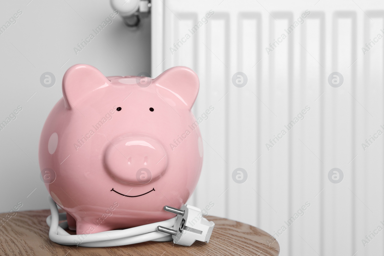 Photo of Piggy bank with plug on wooden table near heating radiator. Space for text