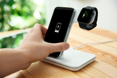 Photo of Man putting mobile phone onto wireless charger at wooden table, closeup. Modern workplace accessory
