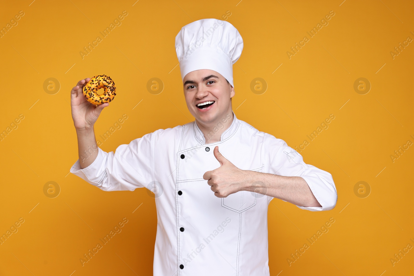 Photo of Portrait of happy confectioner in uniform holding donut on orange background