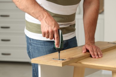 Photo of Man with electric screwdriver assembling furniture at table indoors, closeup