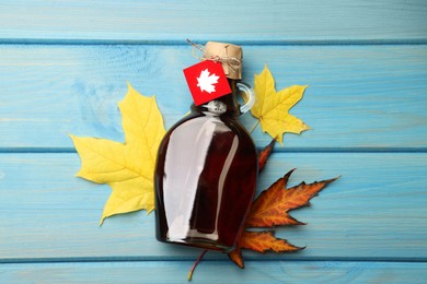 Photo of Glass bottle of tasty maple syrup and dry leaves on light blue wooden table, flat lay