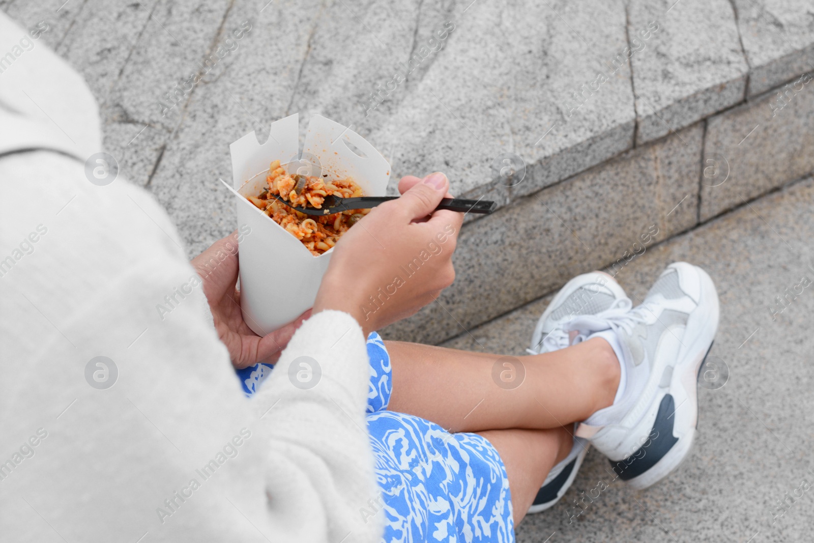 Photo of Woman eating takeaway noodles from paper box with fork outdoors, closeup. Street food