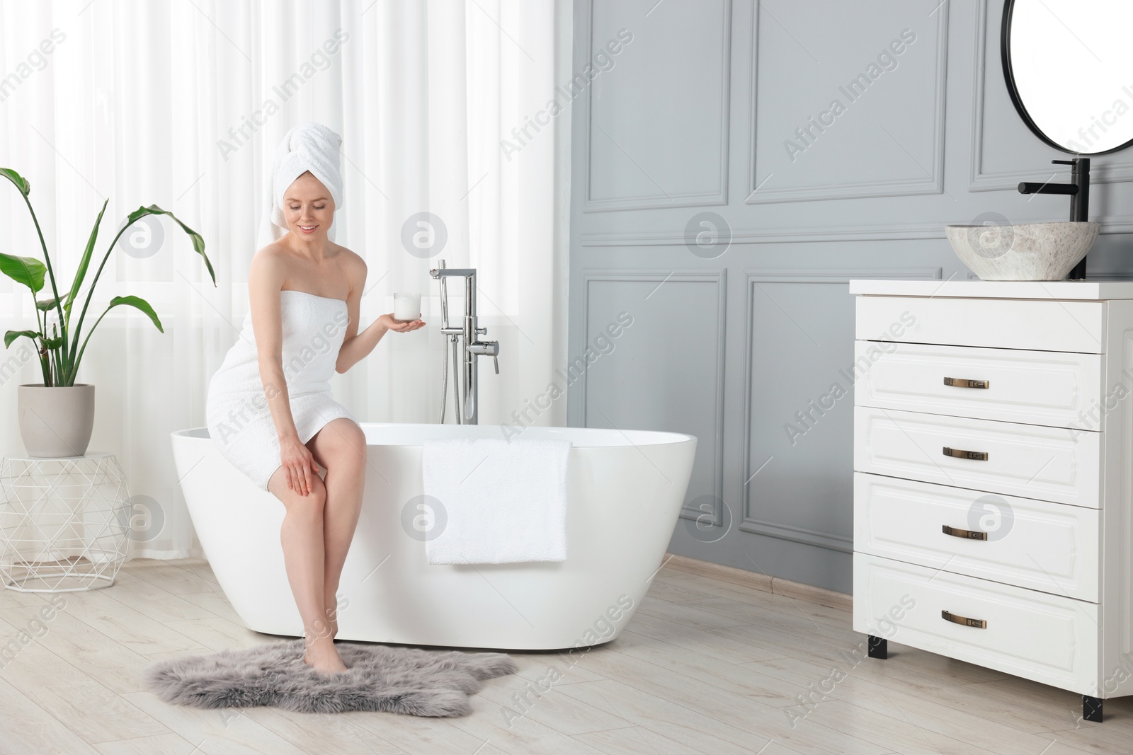 Photo of Beautiful young woman applying body cream onto leg in bathroom