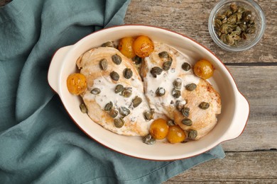 Delicious chicken fillets with capers, tomatoes and sauce in baking dish on wooden table, flat lay