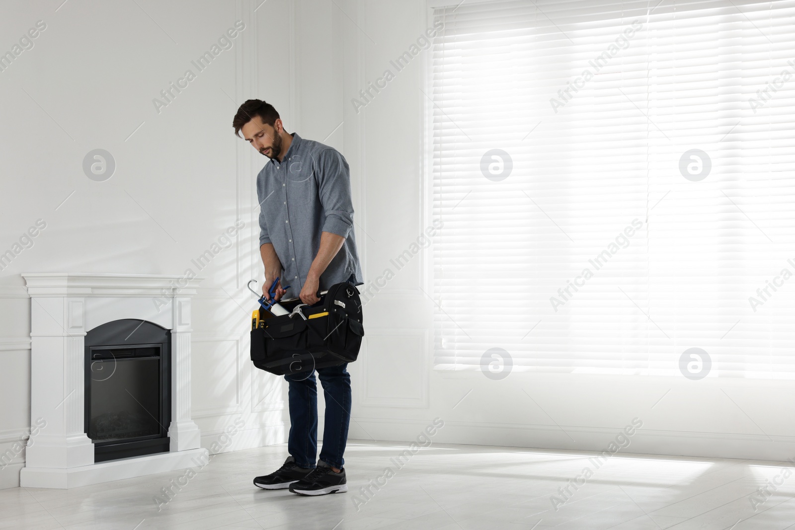Photo of Man with tools installing electric fireplace near white wall in room. Space for text
