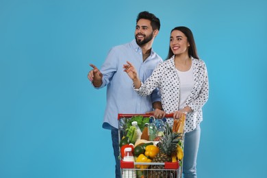 Photo of Young couple with shopping cart full of groceries on light blue background