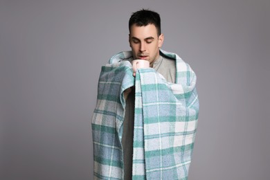 Photo of Young man wrapped in blanket with cup of hot drink suffering from cold on grey background