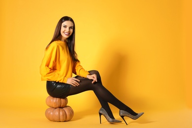 Photo of Beautiful woman sitting on pumpkins against yellow background, space for text. Halloween party