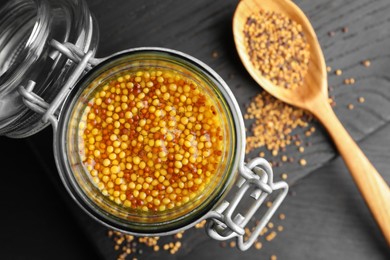Photo of Whole grain mustard in jar and dry seeds on black wooden table, flat lay