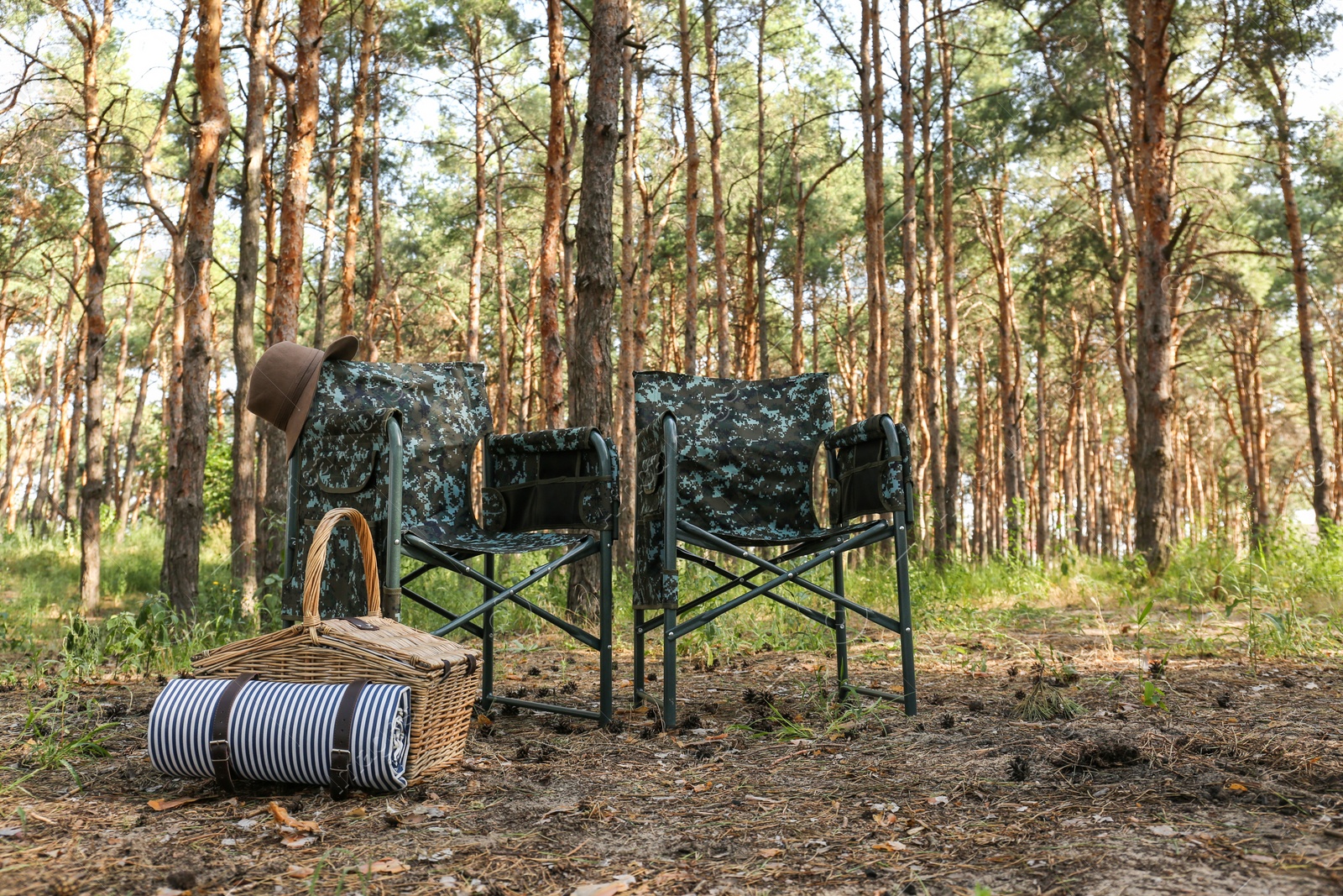 Photo of Camouflage chairs with hat and picnic basket in forest on sunny day