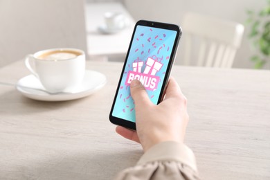 Bonus gaining. Woman using smartphone at white wooden table, closeup. Illustration of gift boxes, word and falling confetti on device screen