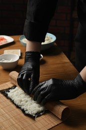 Photo of Chef in gloves making sushi roll at wooden table, closeup