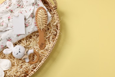 Photo of Different baby accessories, clothes and blank card in wicker basket on yellow background, top view. Space for text