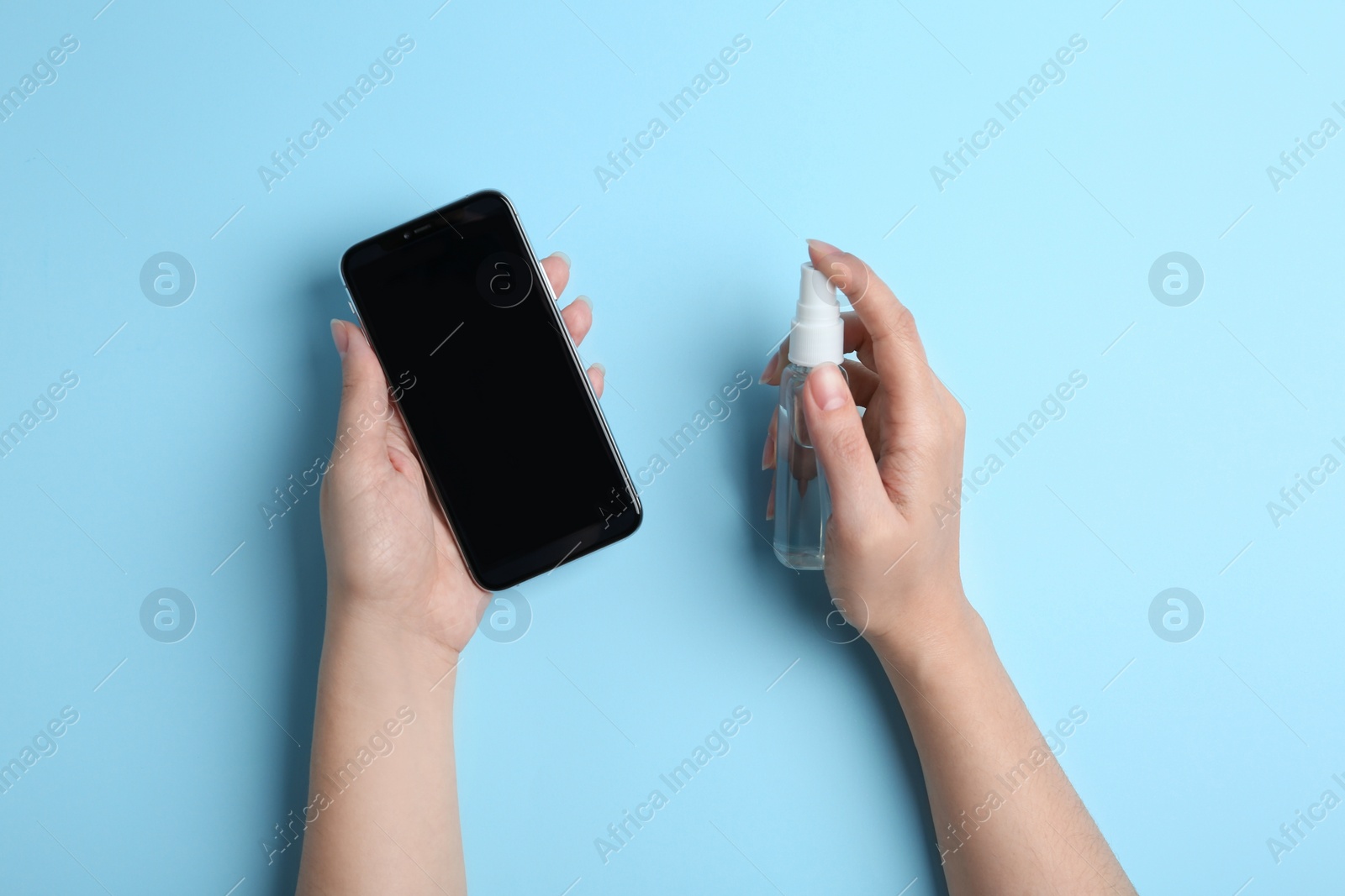 Photo of Woman spraying antiseptic onto smartphone on light blue background, top view