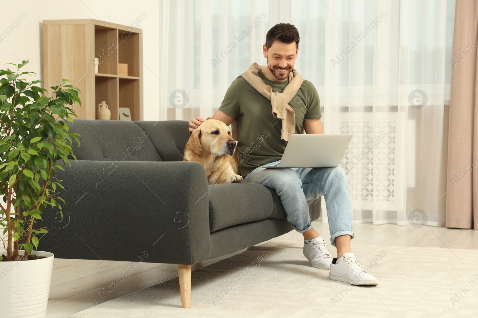 Photo of Man using laptop on sofa near his cute Labrador Retriever at home