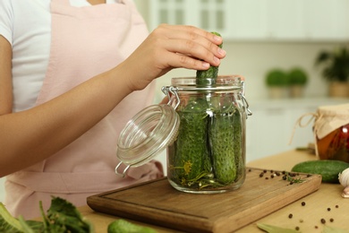 Woman putting cucumber into pickling jar at table in kitchen, closeup