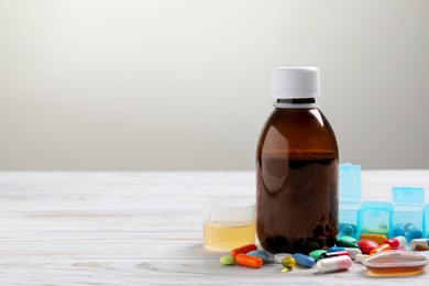 Bottle of syrup, measuring cup, dosing spoon and pills on white wooden table against light grey background, space for text. Cold medicine