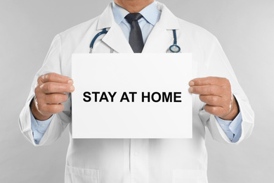 Image of Doctor holding sign with text STAY AT HOME on light grey background, closeup. Quarantine during coronavirus outbreak