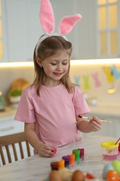 Easter celebration. Cute girl with bunny ears painting eggs at white marble table in kitchen