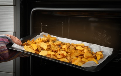 Photo of Woman taking hot roasted potatoes out of oven, closeup