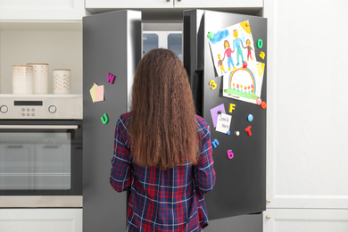 Photo of Woman opening refrigerator door with child's drawings, notes and magnets in kitchen