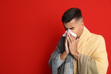 Man wrapped in warm blanket sneezing on red background, space for text. Cold symptoms