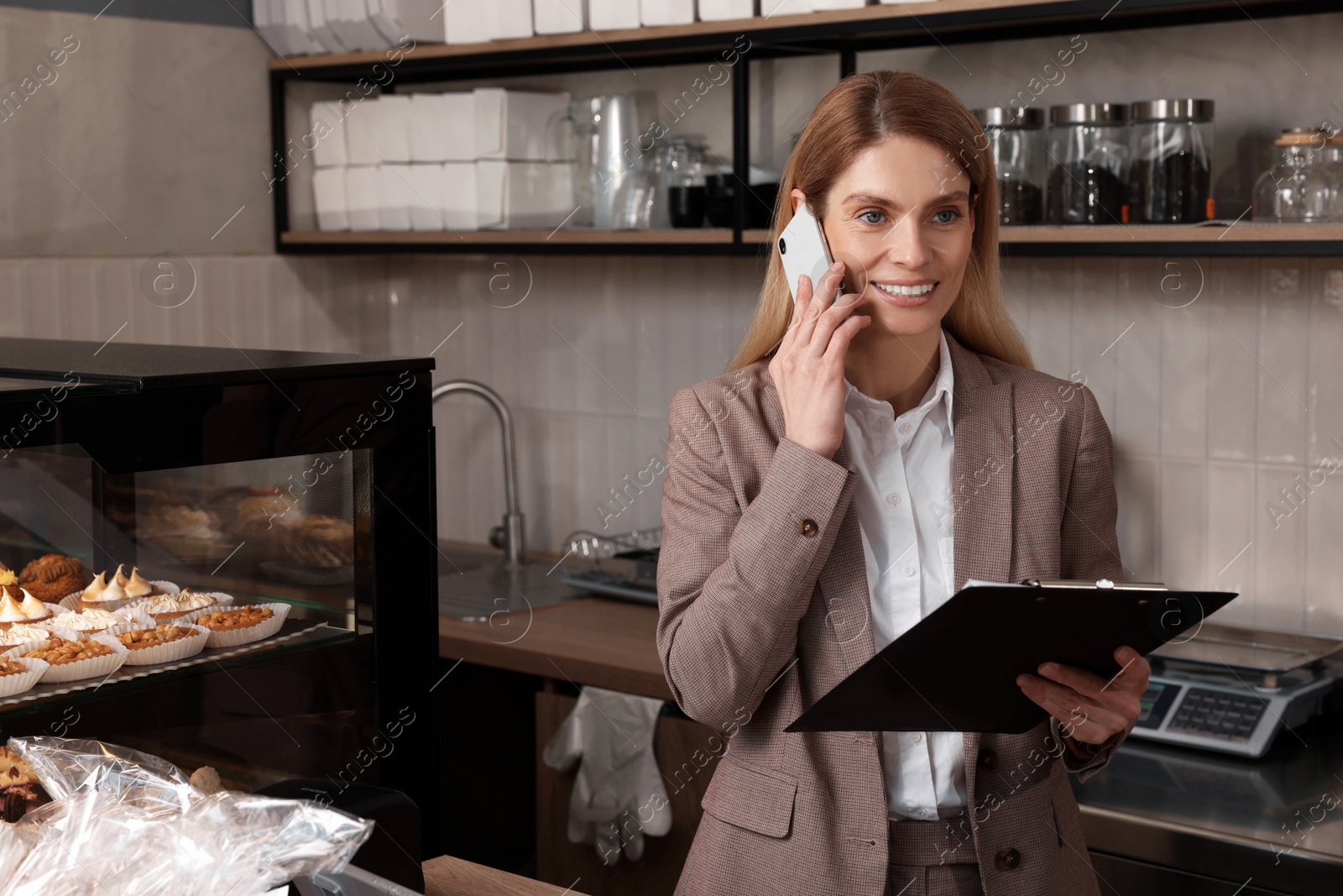 Photo of Happy business owner with clipboard talking on phone in bakery shop