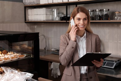 Photo of Happy business owner with clipboard talking on phone in bakery shop