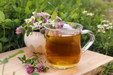 Photo of Cup of aromatic herbal tea and ceramic mortar with different wildflowers on wooden board in meadow