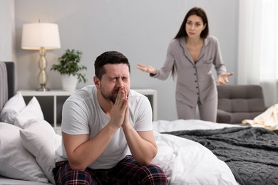 Photo of Stressed husband sitting on bed while his wife blaming him in bedroom, selective focus. Relationship problems