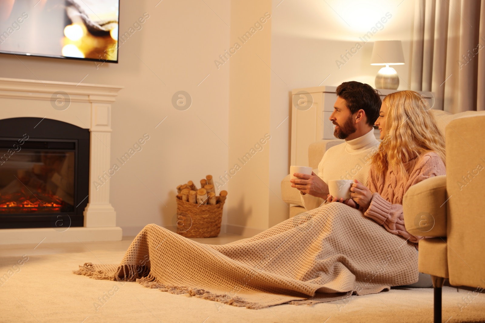 Photo of Lovely couple with hot drinks spending time together near fireplace at home