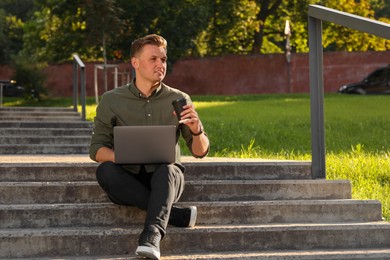 Handsome man with cup of coffee and laptop on stairs outdoors. Space for text