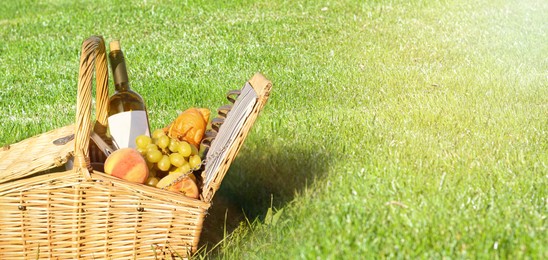 Image of Picnic basket with snacks and bottle of wine on green grass in park, space for text. Banner design