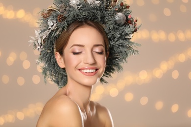 Beautiful young woman wearing Christmas wreath against blurred festive lights. Space for text