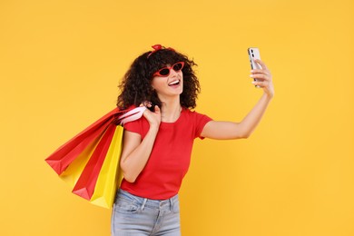 Photo of Happy young woman with shopping bags taking selfie on yellow background