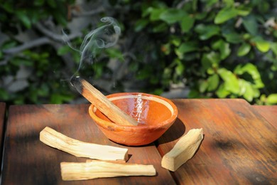 Photo of Palo santo stick smoldering in bowl on wooden table outdoors