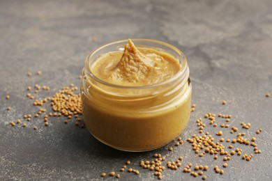 Photo of Tasty mustard sauce in jar and dry seeds on grey textured table, closeup