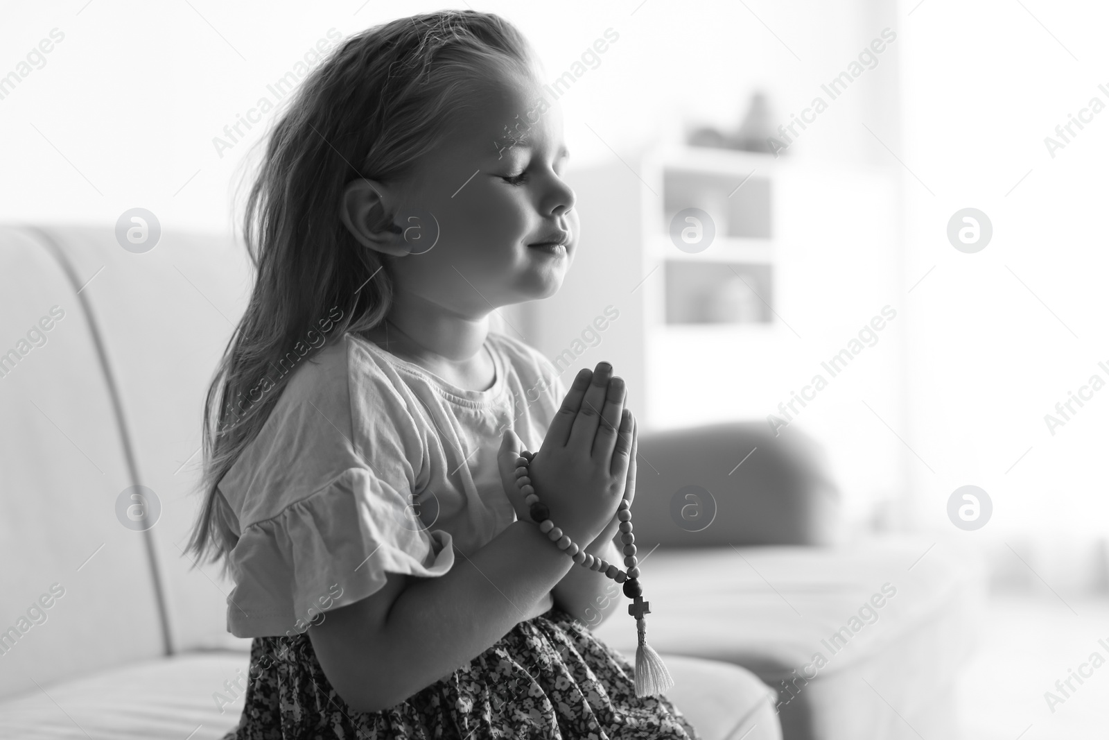 Photo of Cute little girl with beads praying on sofa in living room, black and white effect. Space for text