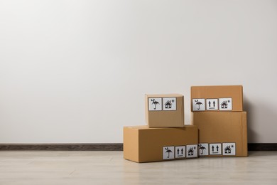 Photo of Many closed cardboard boxes with packaging symbols on floor near white wall, space for text. Delivery service
