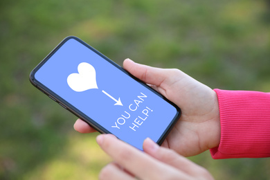 Image of Donations concept. Woman holding smartphone with text YOU CAN HELP outdoors, closeup