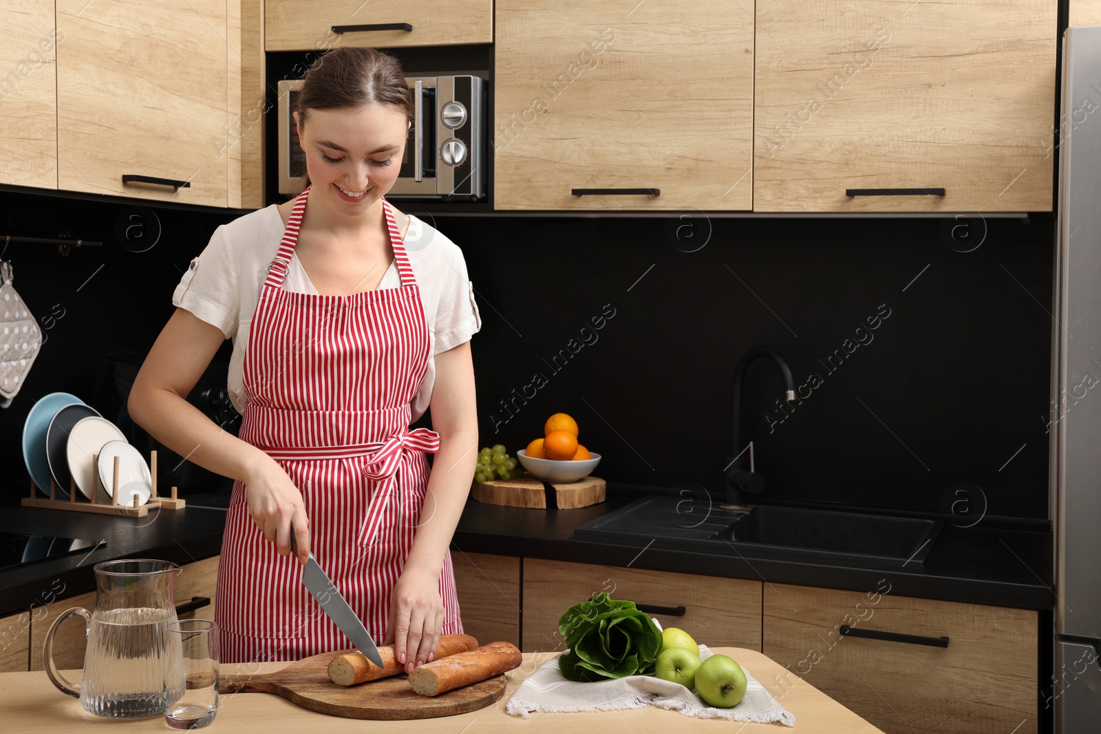 Photo of Young woman in striped apron cutting baguette on wooden table at kitchen